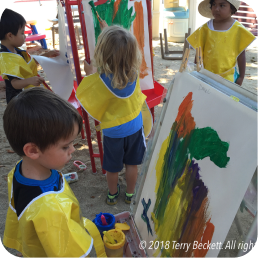 Children painting on easels outdoors in sunlight.